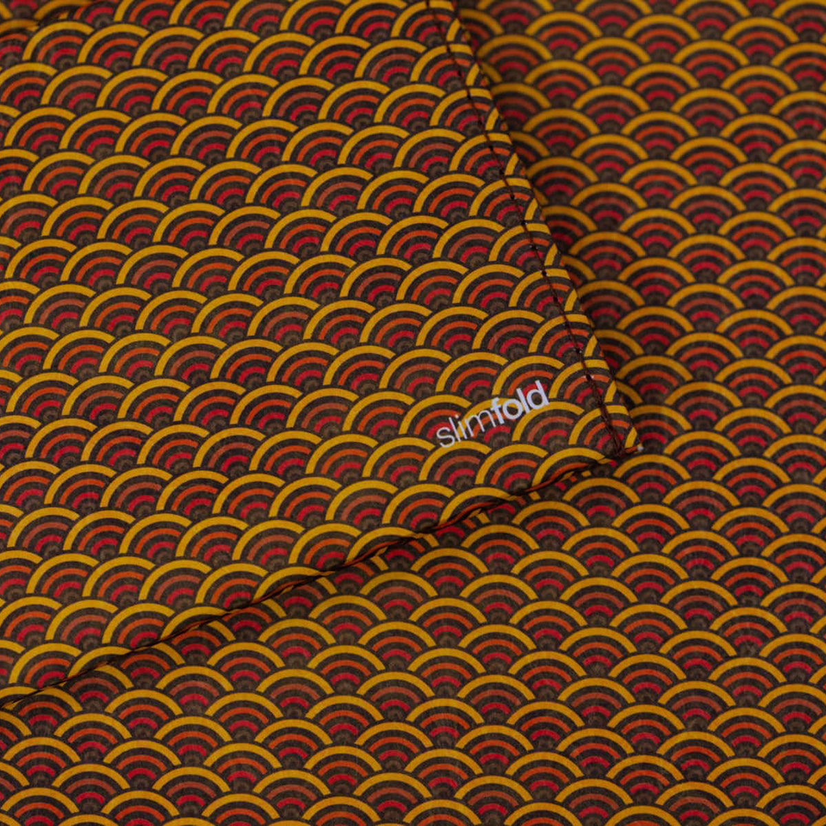 A close-up of a SlimFold Micro Tyvek wallet with a repeating Japanese-style wave pattern in mustard yellow, red, and dark brown, with a small white 'simfold' logo printed in the center.